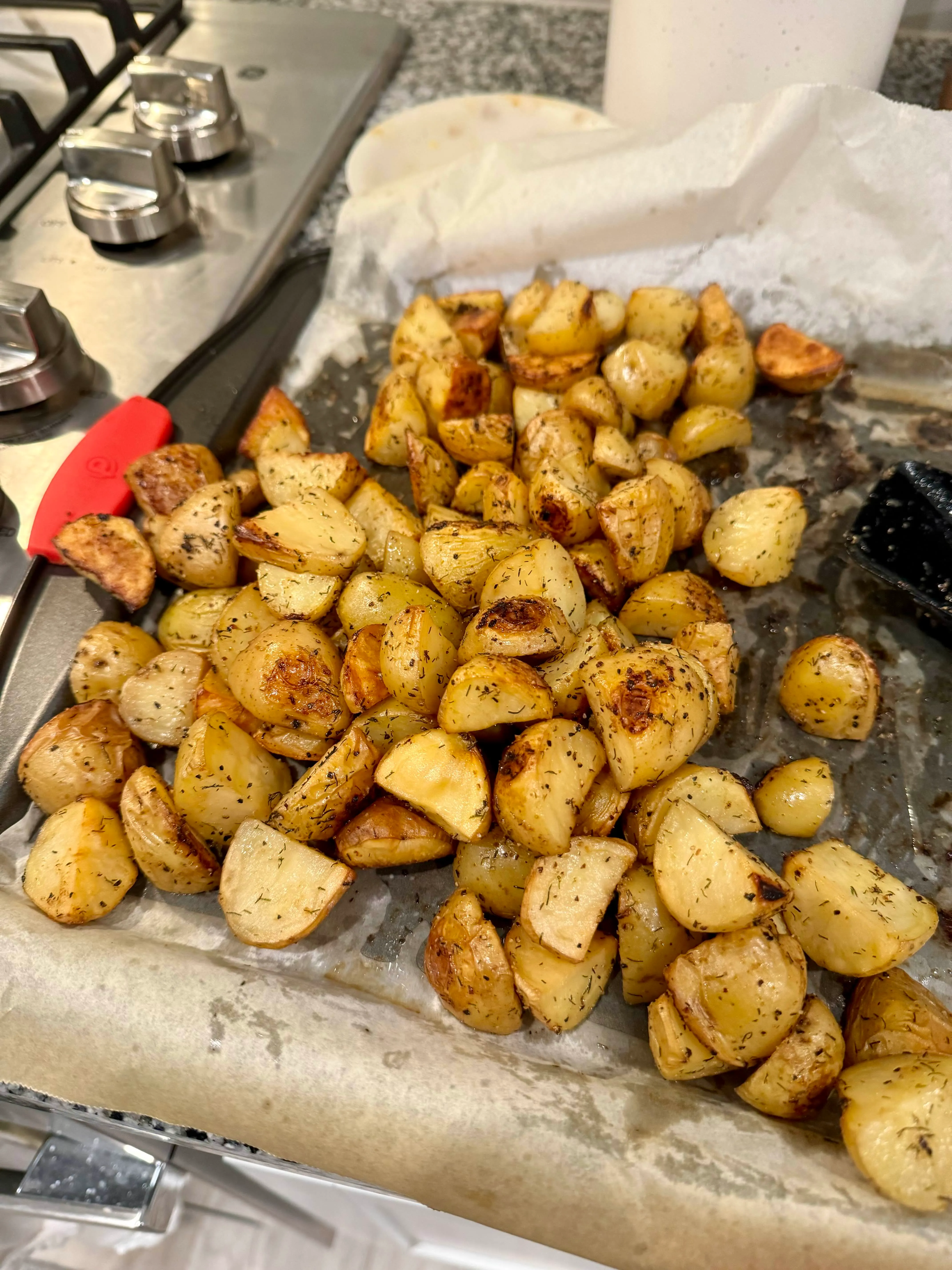 A sheet pan crowded with golden roast potato chunks, dried herbs clinging to every edge.
