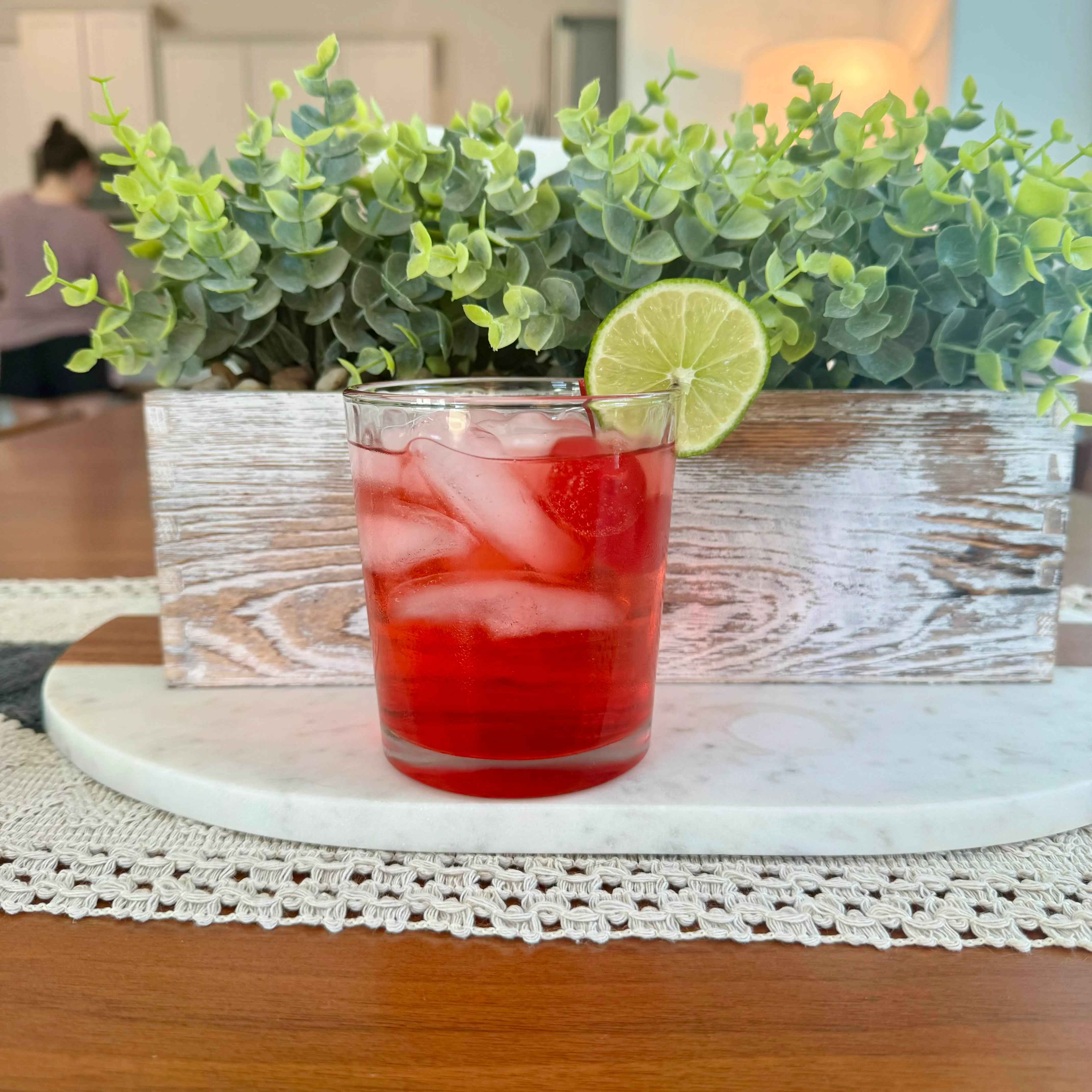 A short rocks glass of ruby-red drink with a lime wheel, set on a marble board against eucalyptus.