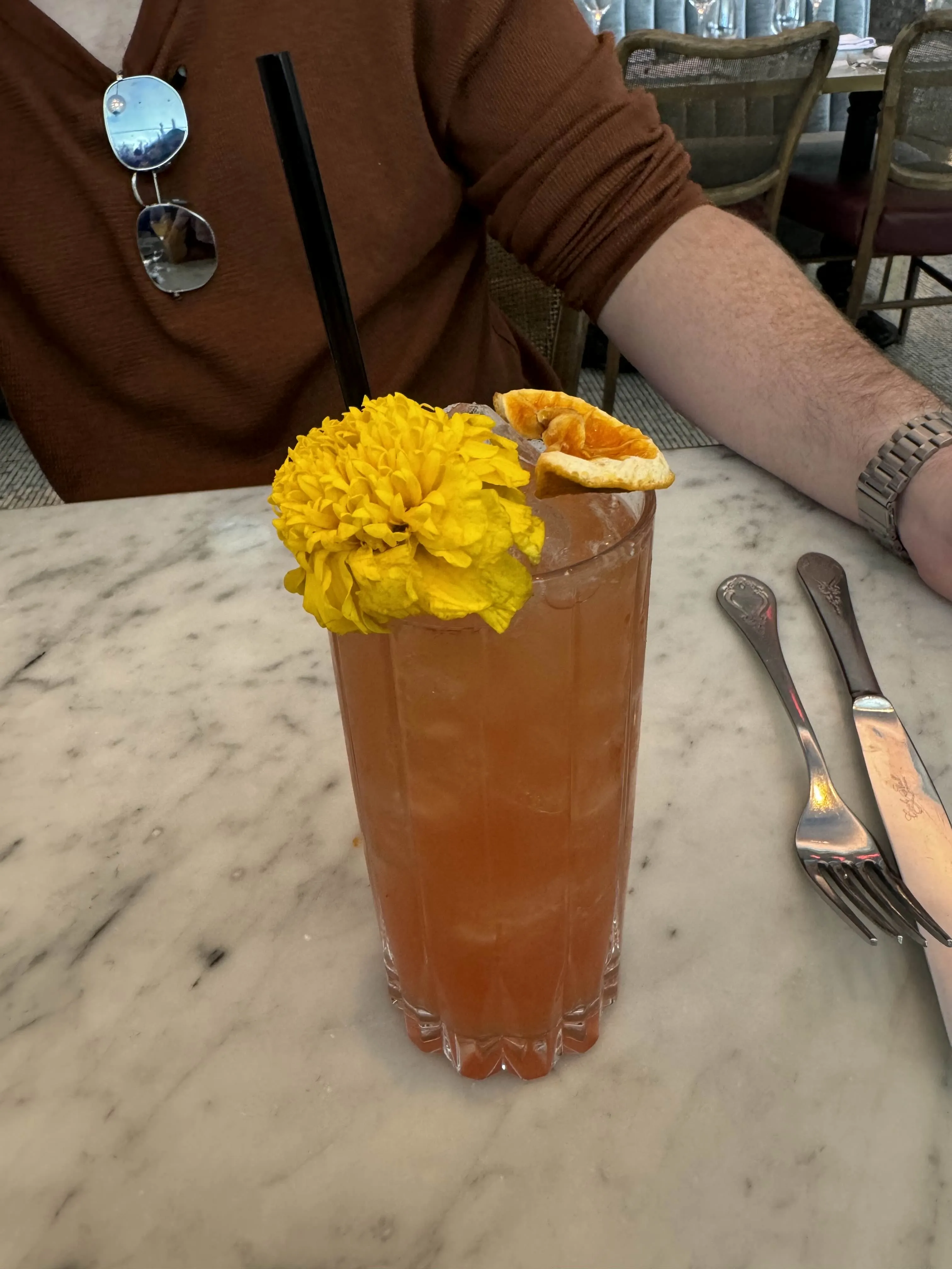 A tall cocktail on a marble table, garnished with a marigold flower and a dried orange slice.