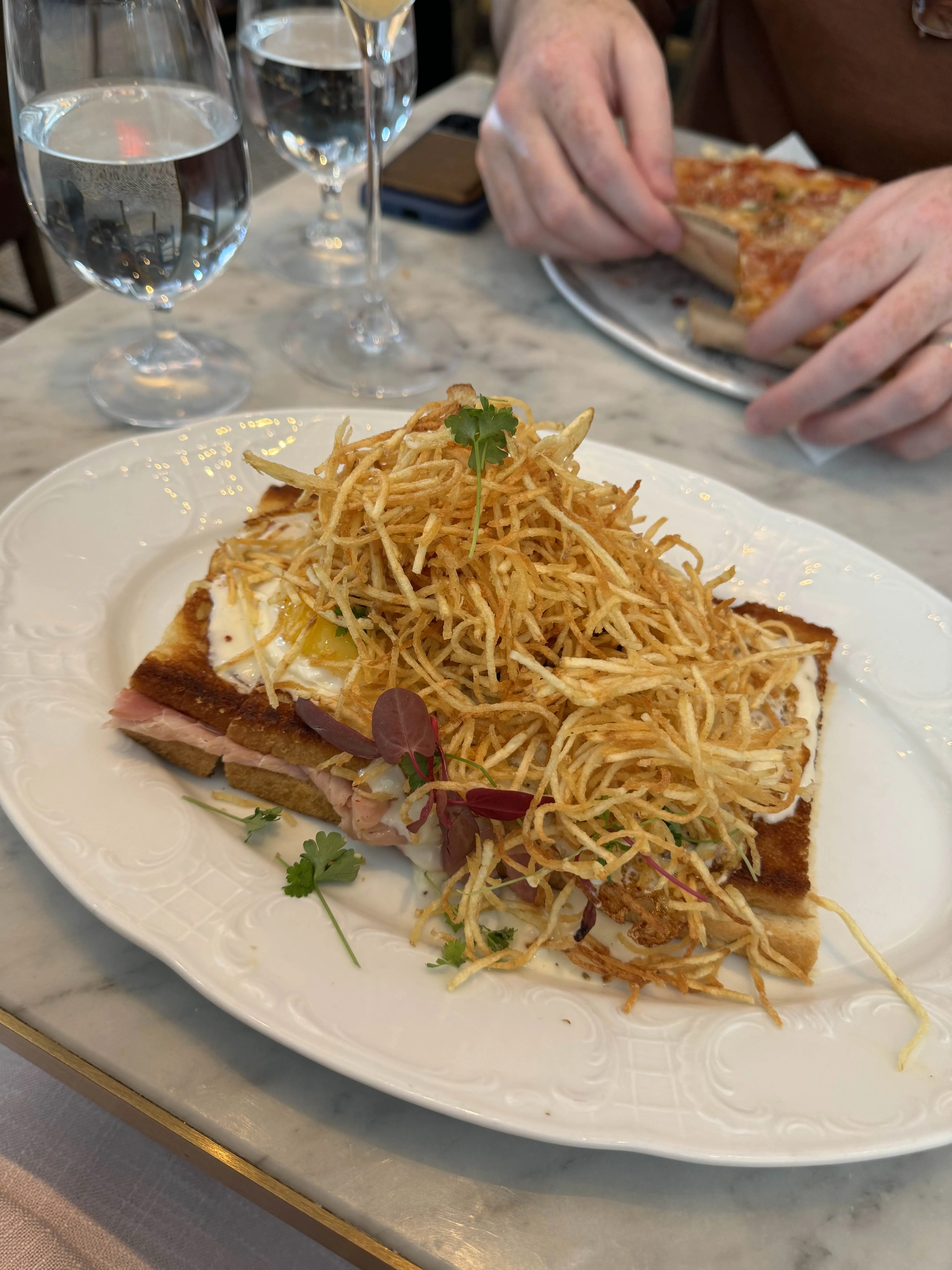 An open-faced sandwich with pink ham and a fried egg, heaped with a tangle of shoestring fries, on a marble tabletop.