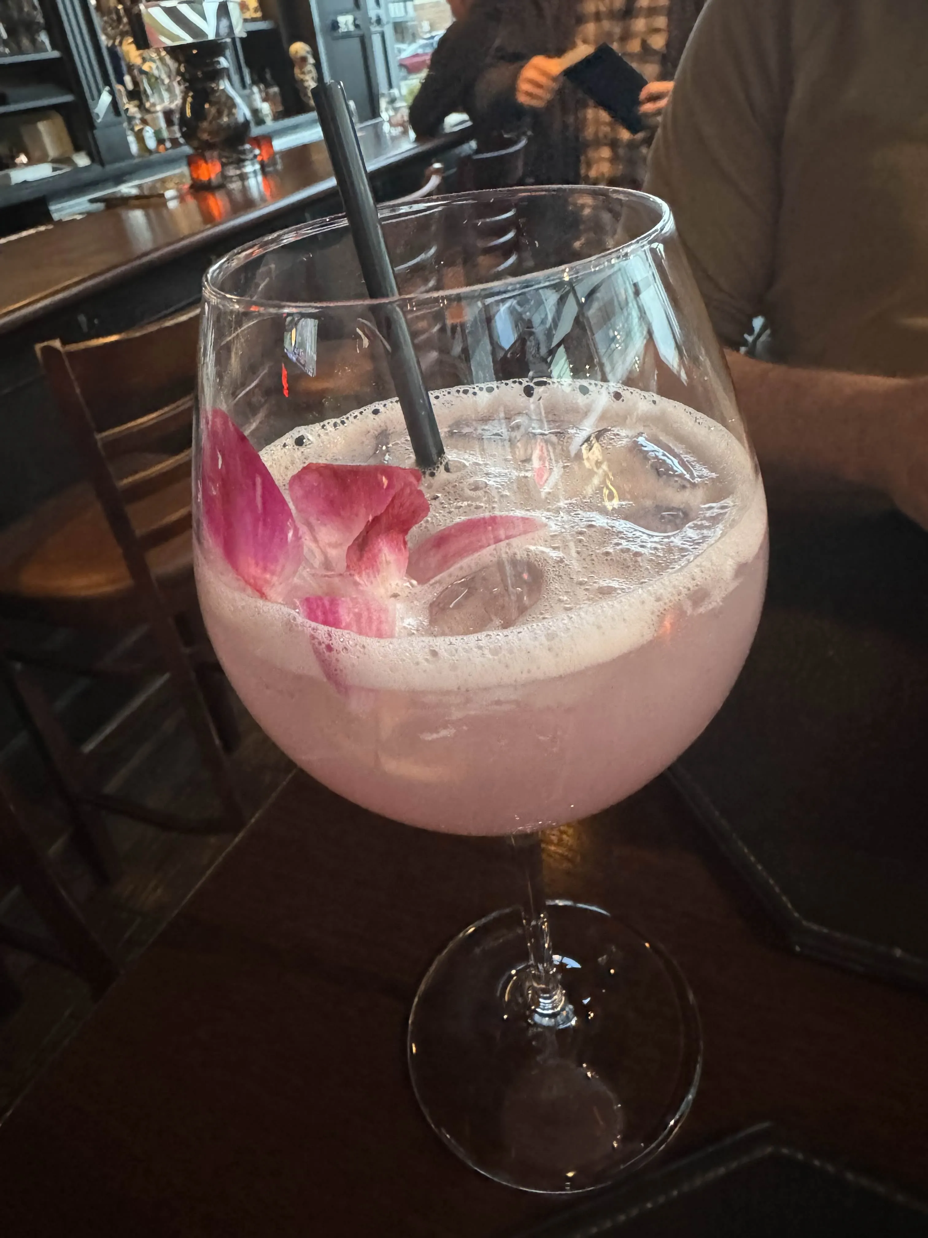 A large coupe of pale-pink cocktail with rose petals floating on top and a single black straw, on a dark wooden bar top.
