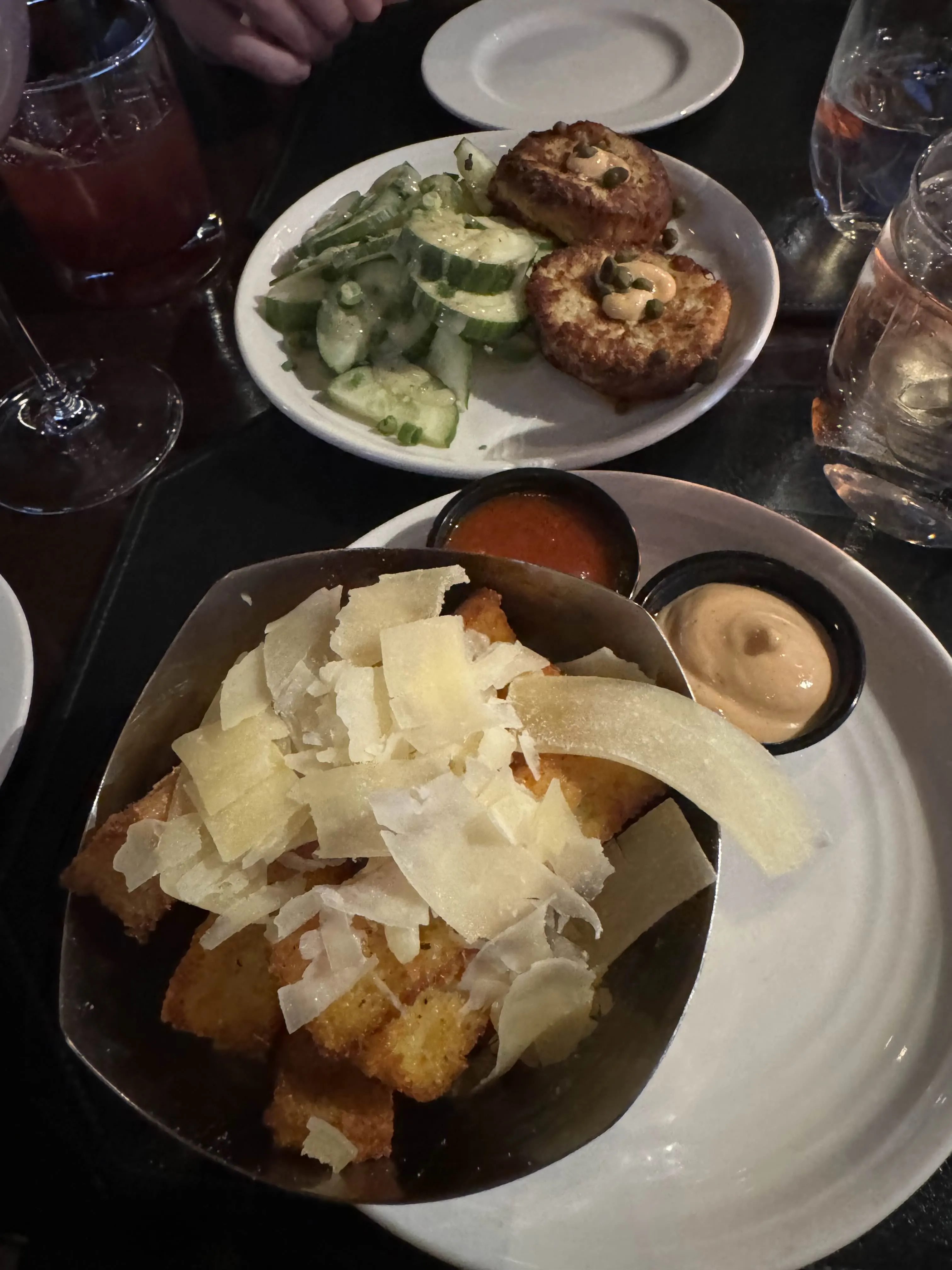 A dark bowl of fried potato wedges showered in shaved parmesan, with two small ramekins of red sauce and a peach-colored aioli.