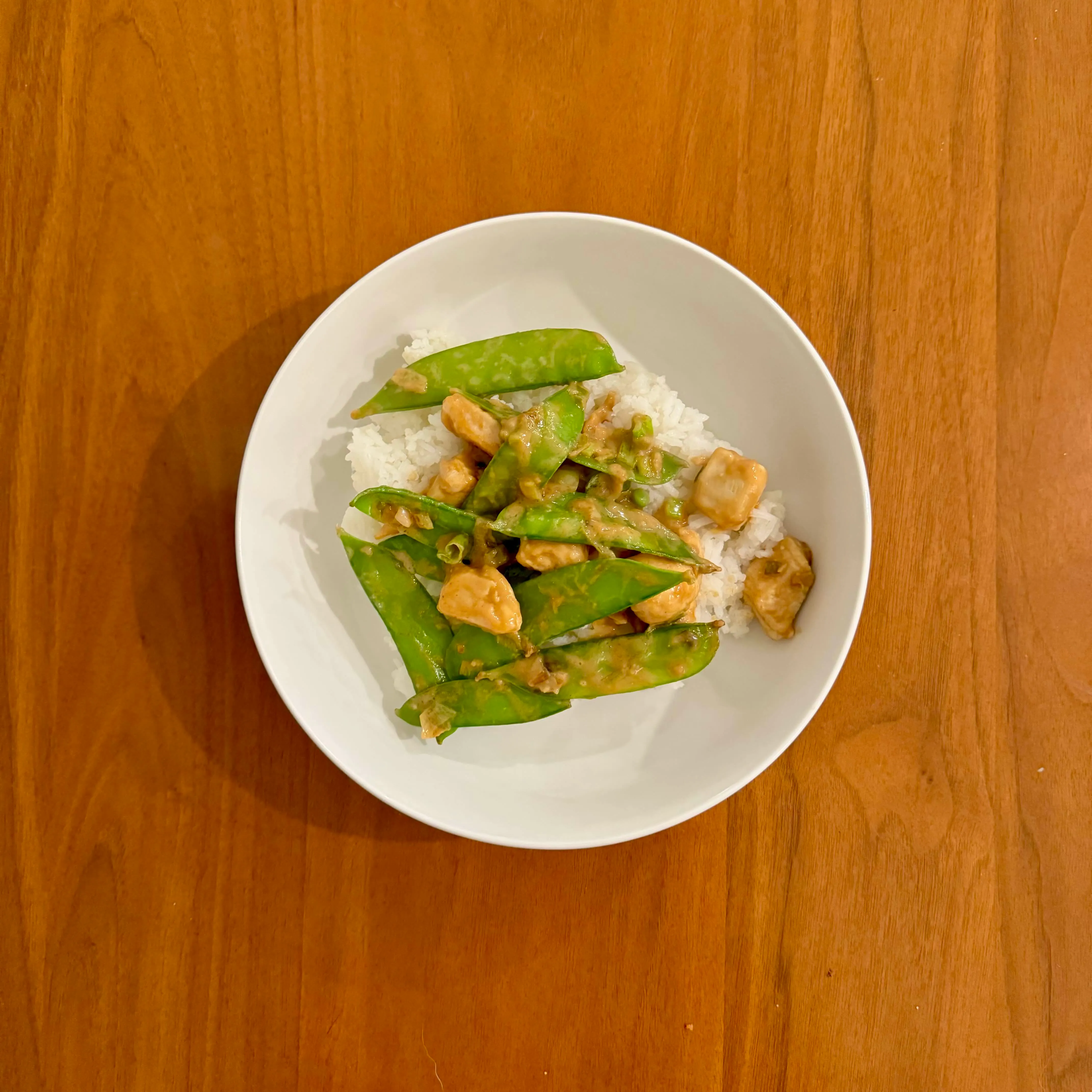 A shallow bowl of white rice topped with a snow-pea and chicken stir fry in a glossy peanut-colored sauce.