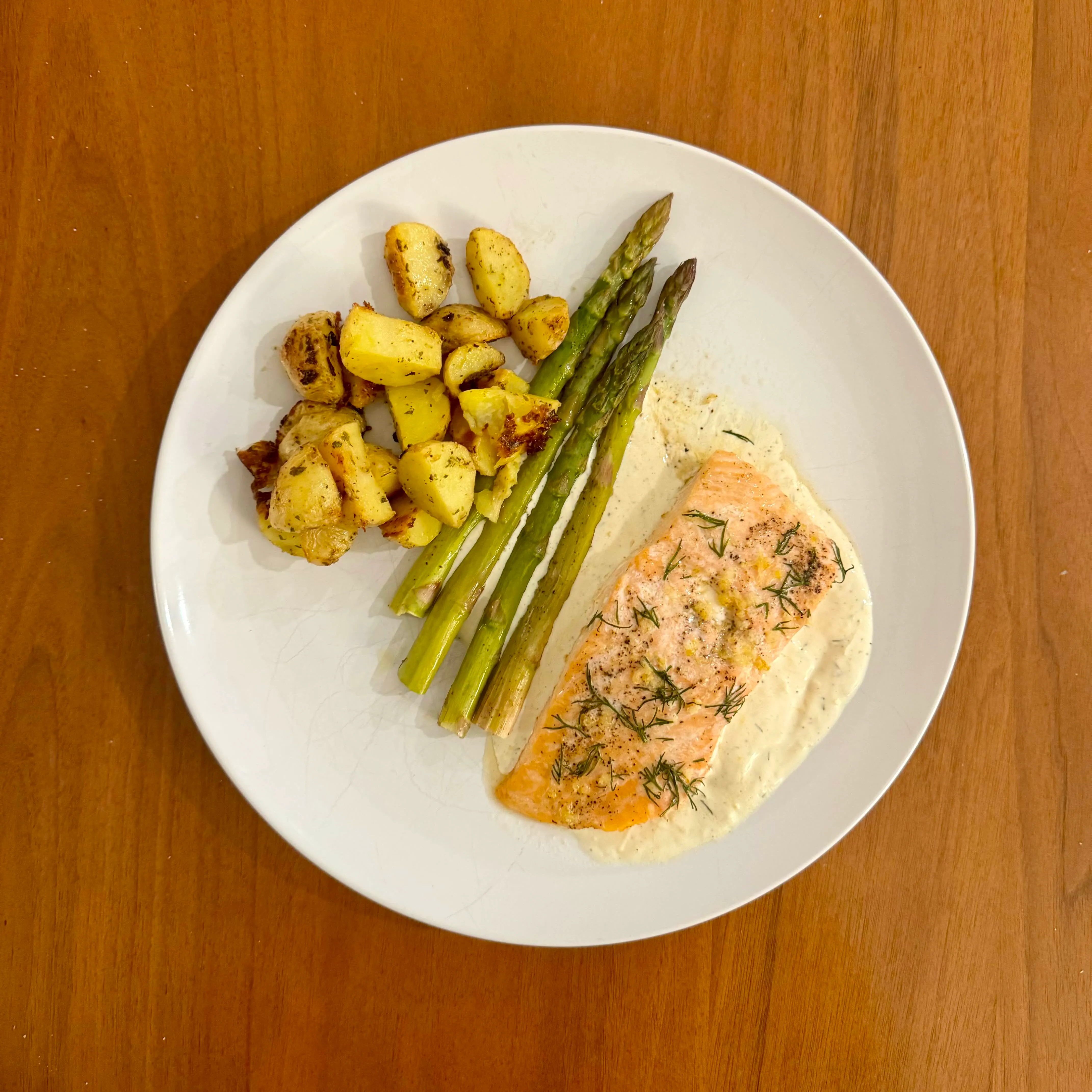 A pink salmon fillet with dill on a pool of cream sauce, beside roasted potato chunks and tall green asparagus spears.