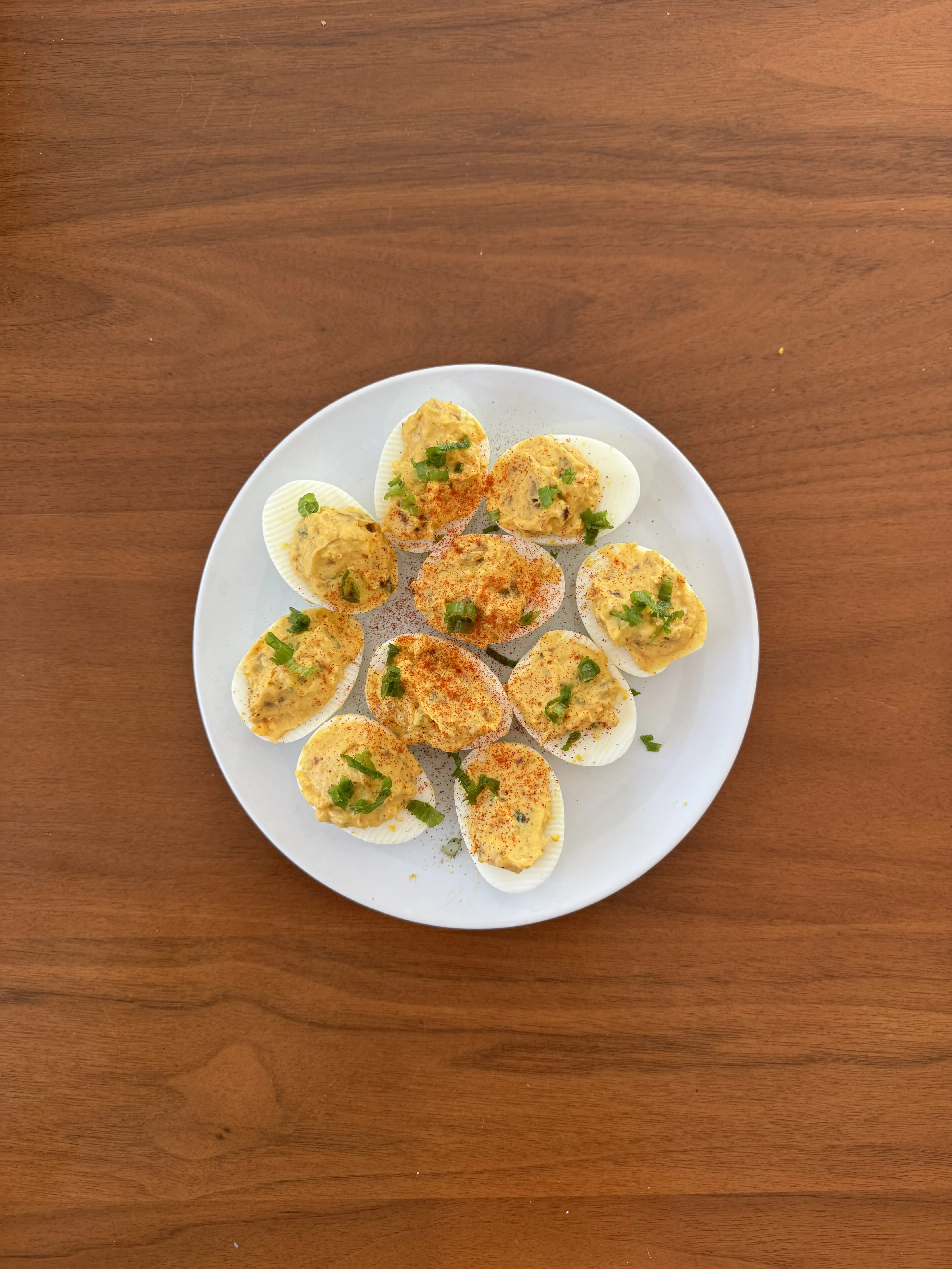 A plate of deviled eggs arranged in a ring, dusted with paprika and sprinkled with chopped chives, on a walnut table.