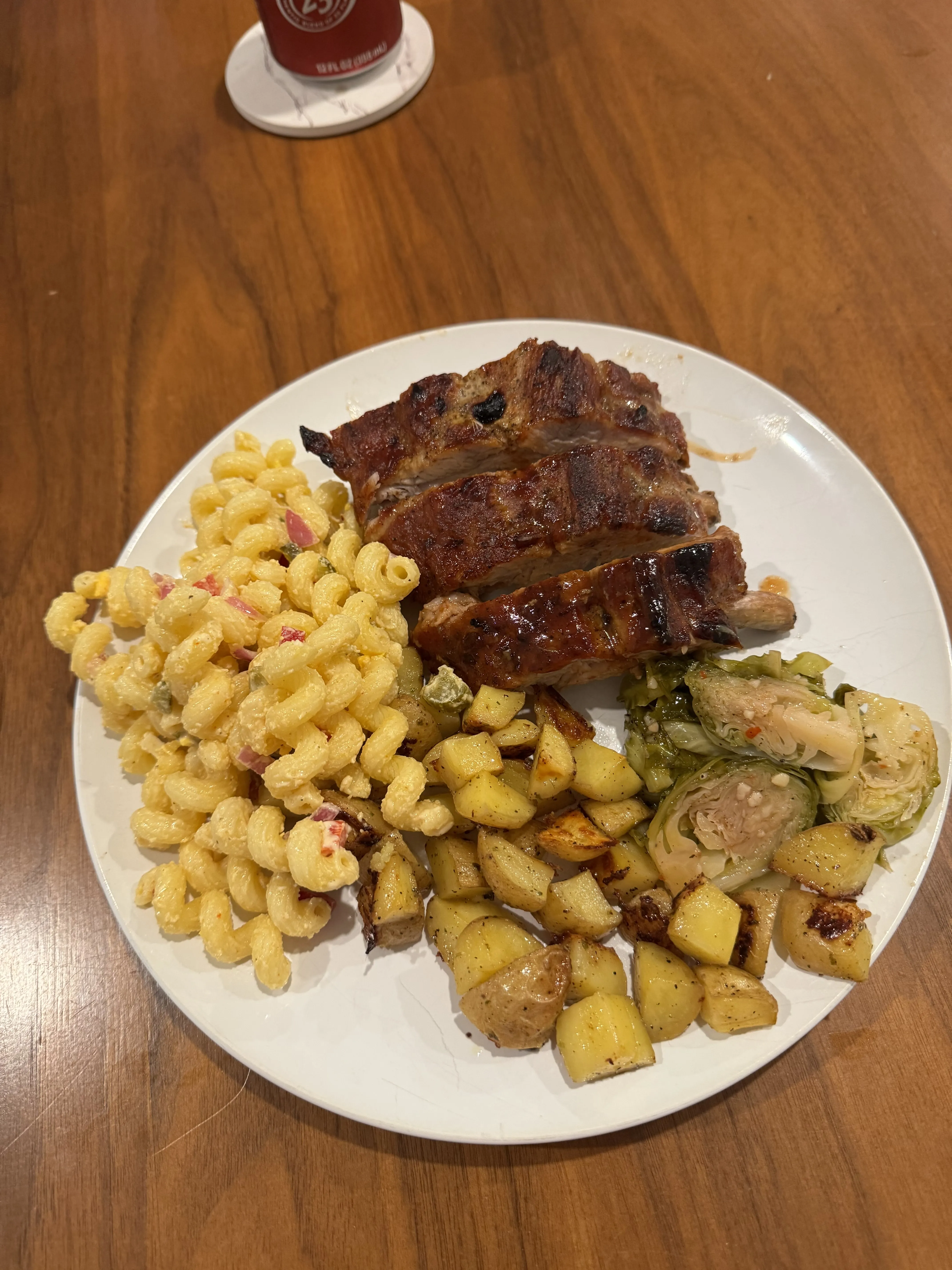 A dinner plate with sliced meatloaf, cold macaroni salad, roast potato cubes, and halved brussels sprouts, a can of beer behind it.