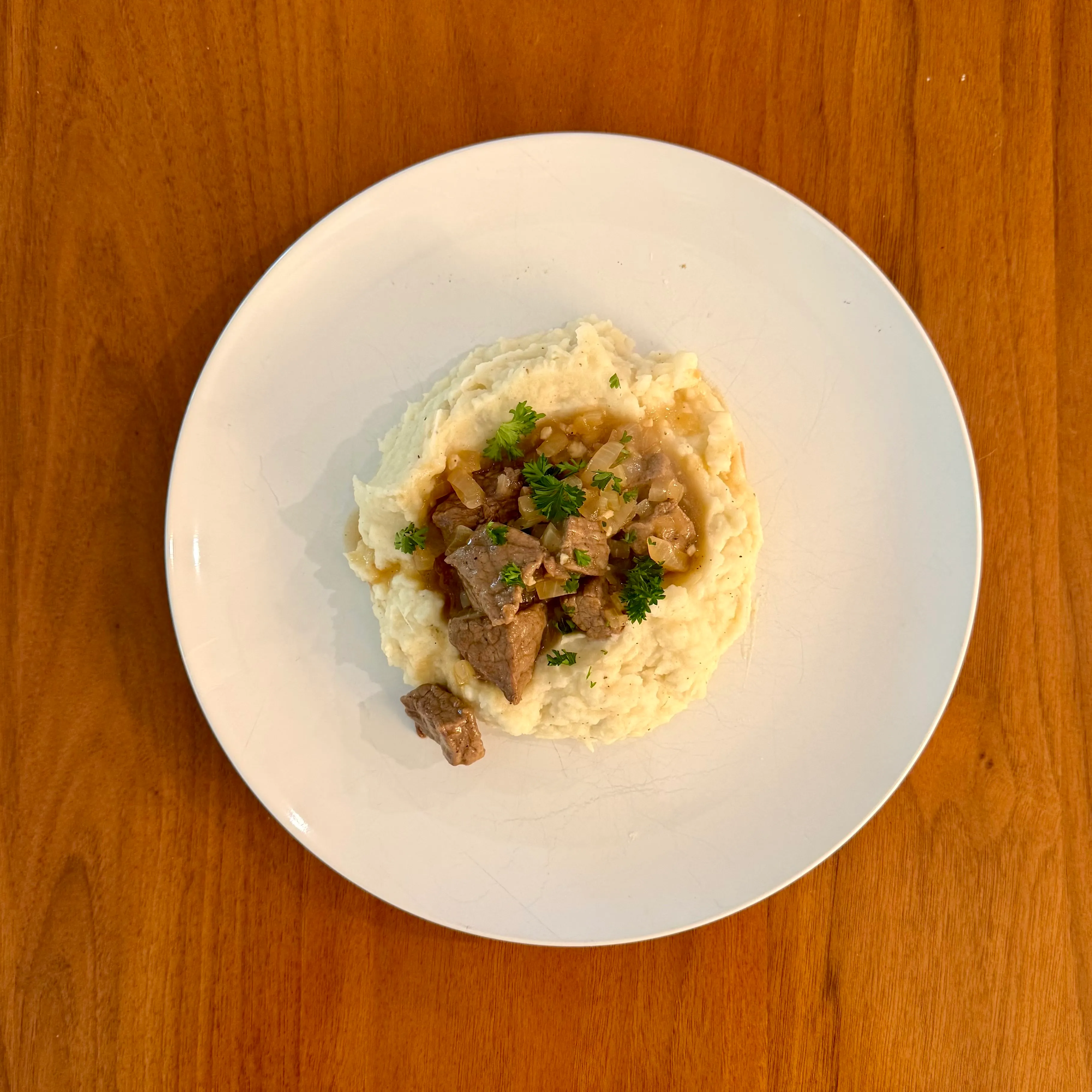 A shallow bowl of mashed potatoes with chunks of beef in a pan gravy piled in the middle, finished with torn parsley.