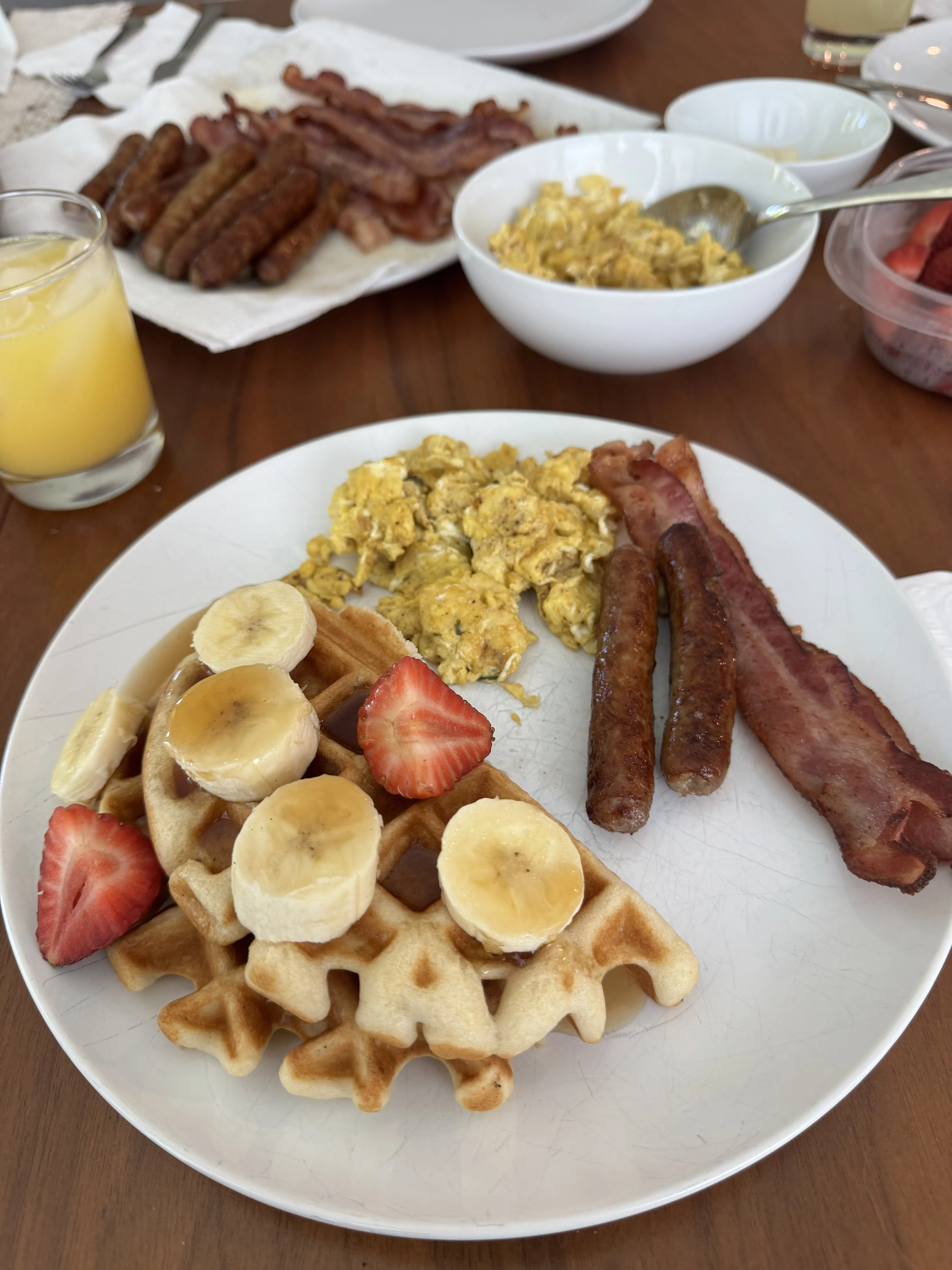 A breakfast plate with a quartered Belgian waffle topped with banana and strawberry, scrambled eggs, and thick bacon. Behind it, a platter of sausages and more bacon, a bowl of eggs, orange juice, and cut strawberries.