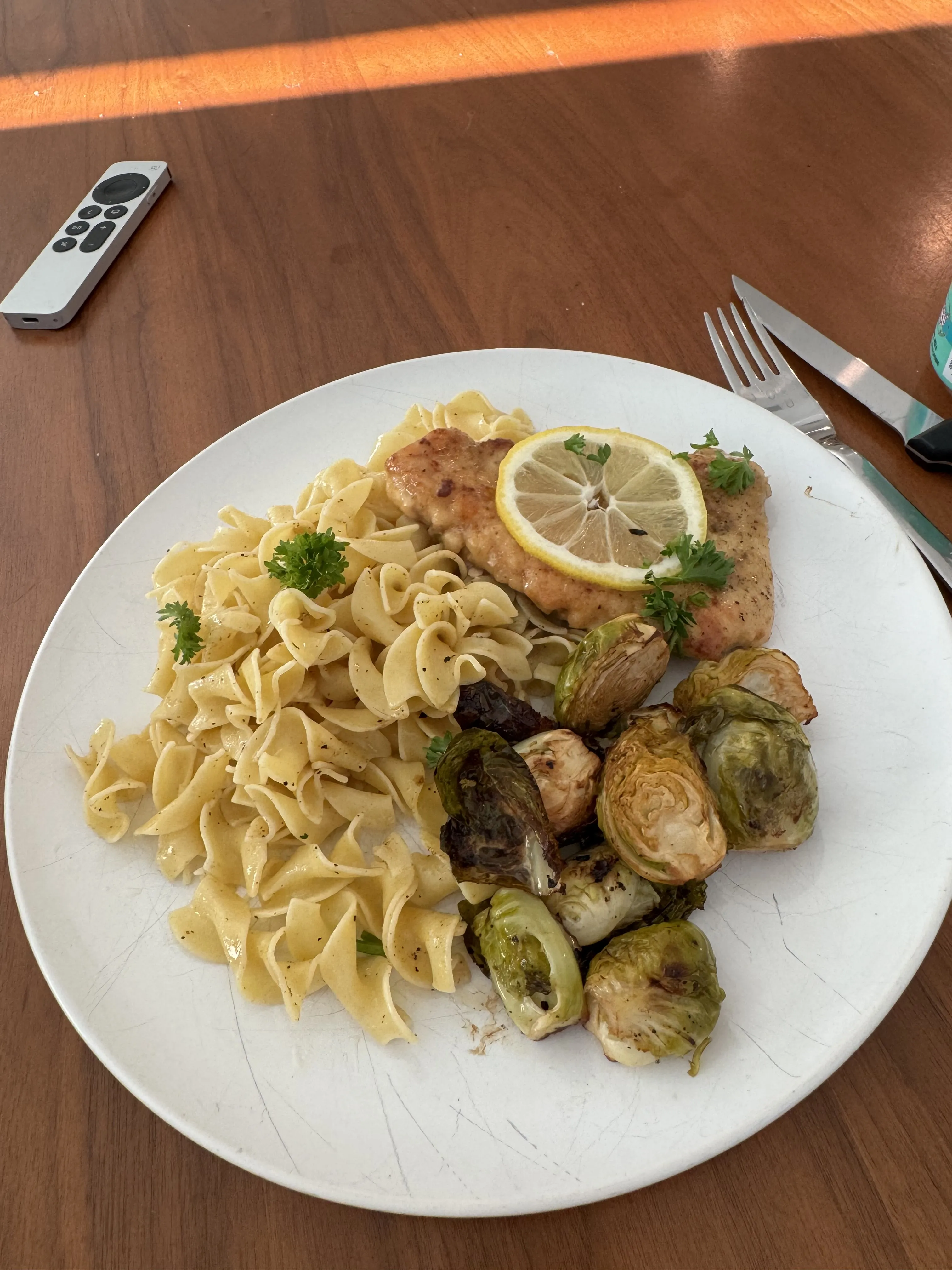 A plate with chicken piccata in lemon-caper sauce, a mound of buttered egg noodles, and roasted brussels sprouts, with a lemon slice resting on the chicken.