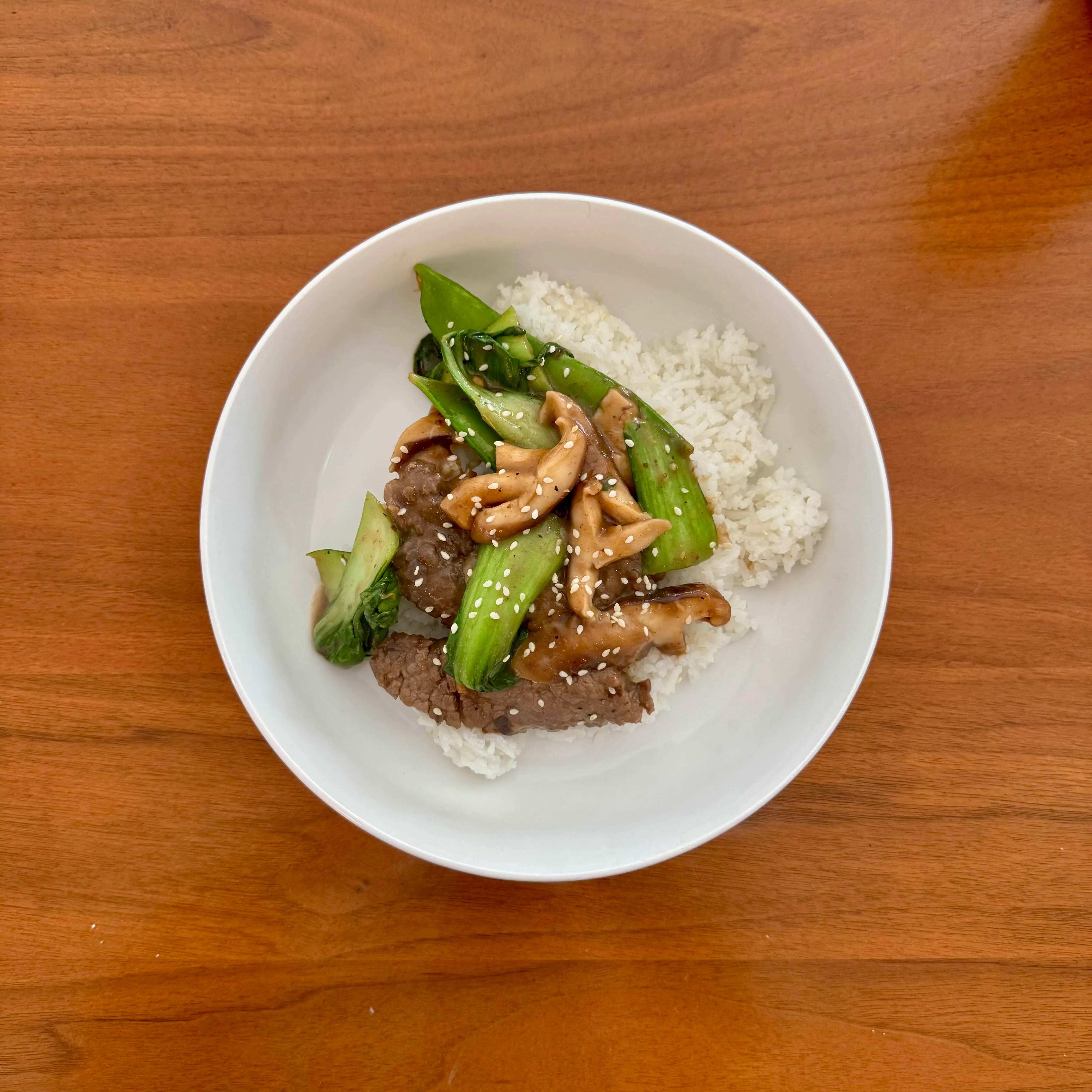 A bowl of mongolian beef with bok choy and mushrooms over white rice, finished with sesame seeds.