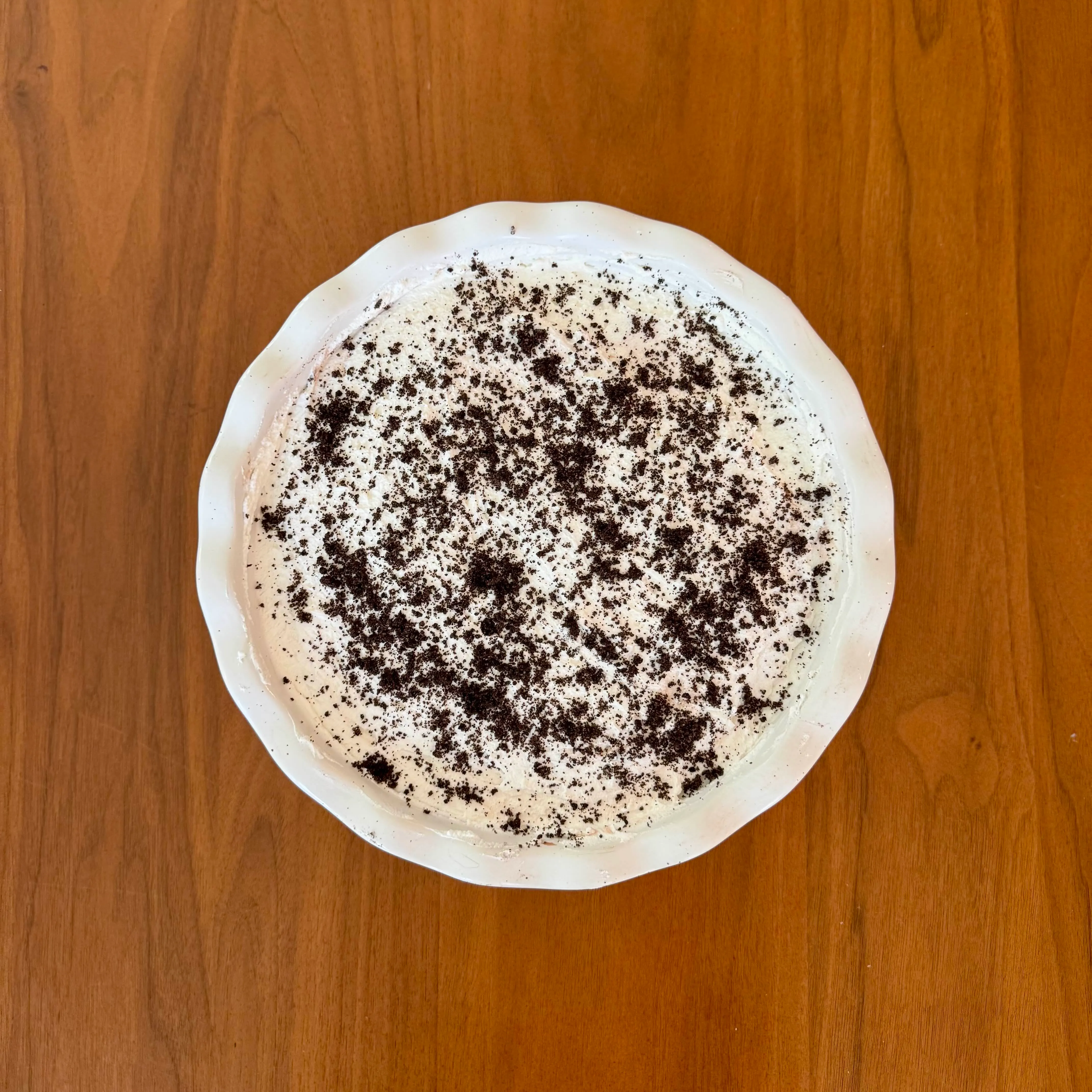 A pale icebox pie in a white fluted dish, covered in whipped topping and heavily dusted with dark cookie crumbs.