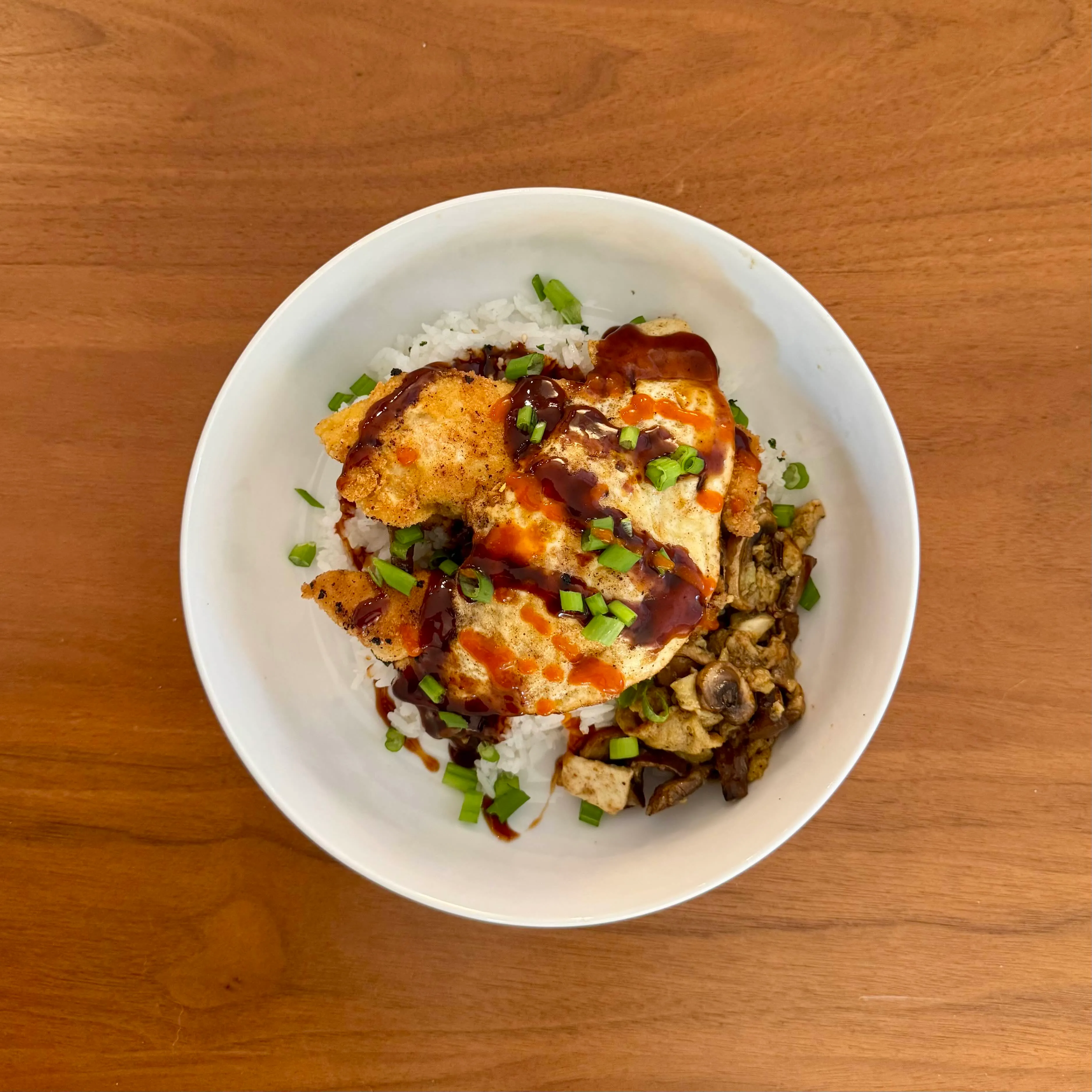 A rice bowl with a crispy chicken cutlet, a fried egg, sliced mushrooms, and drizzles of dark sauce and bright sriracha, finished with green onions.