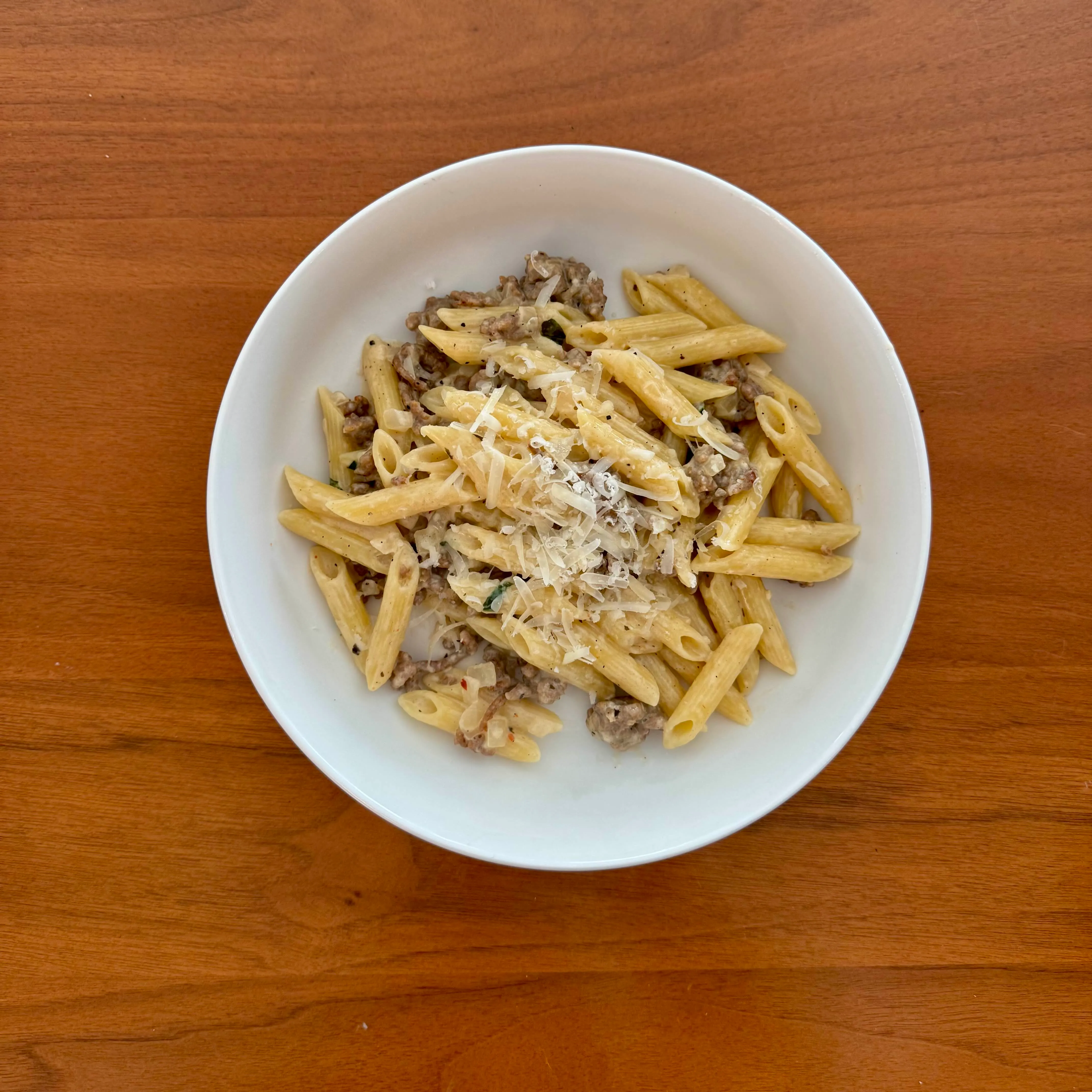 A bowl of penne pasta tossed with crumbled browned meat in a pale cream-and-parmesan sauce, finished with cracked pepper.