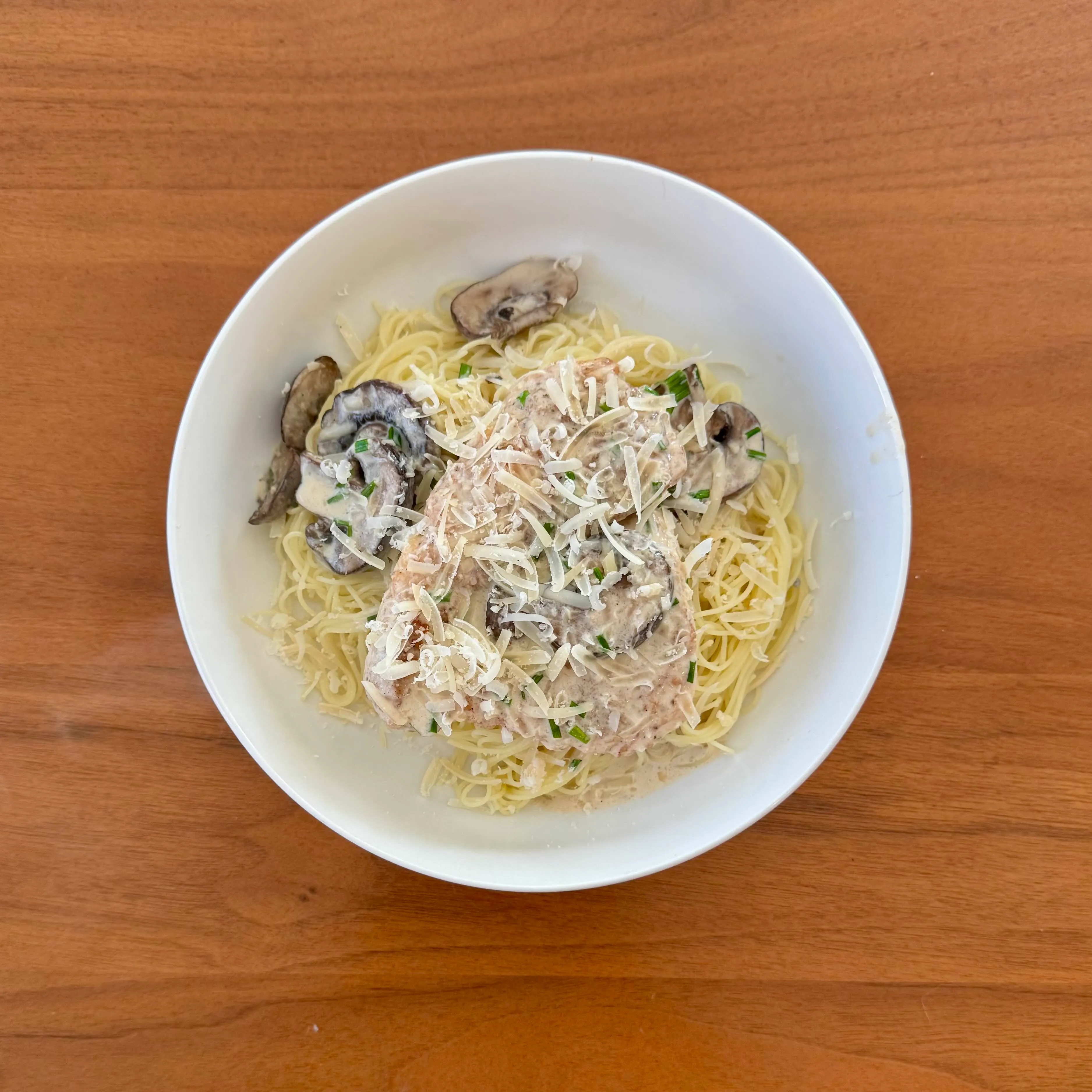A bowl of angel-hair pasta under a blanket of chicken in a mushroom cream sauce with shaved parmesan and chopped chives.