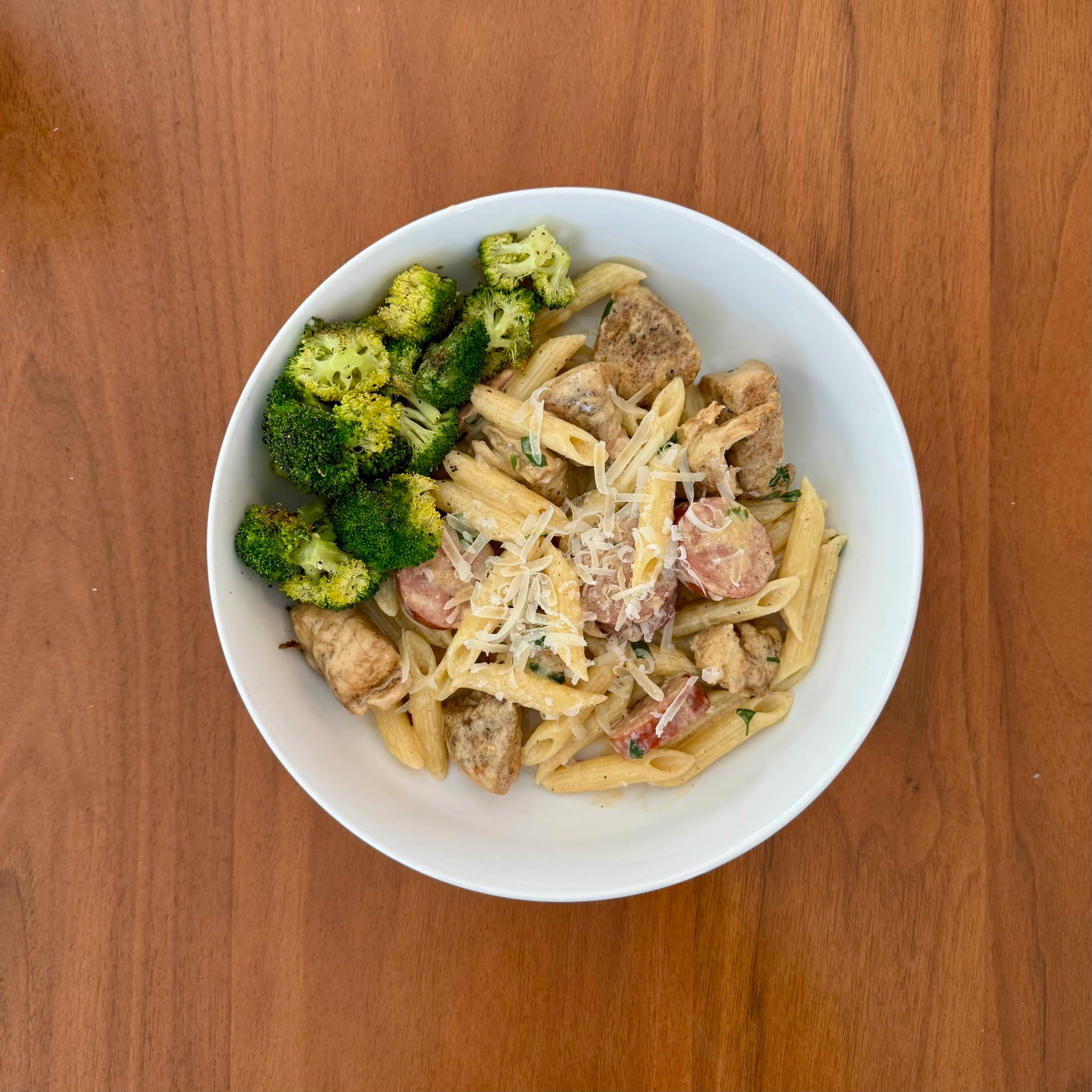 A bowl of penne with pieces of chicken and sausage in a creamy sauce, shaved parmesan on top, with charred broccoli florets on the side.