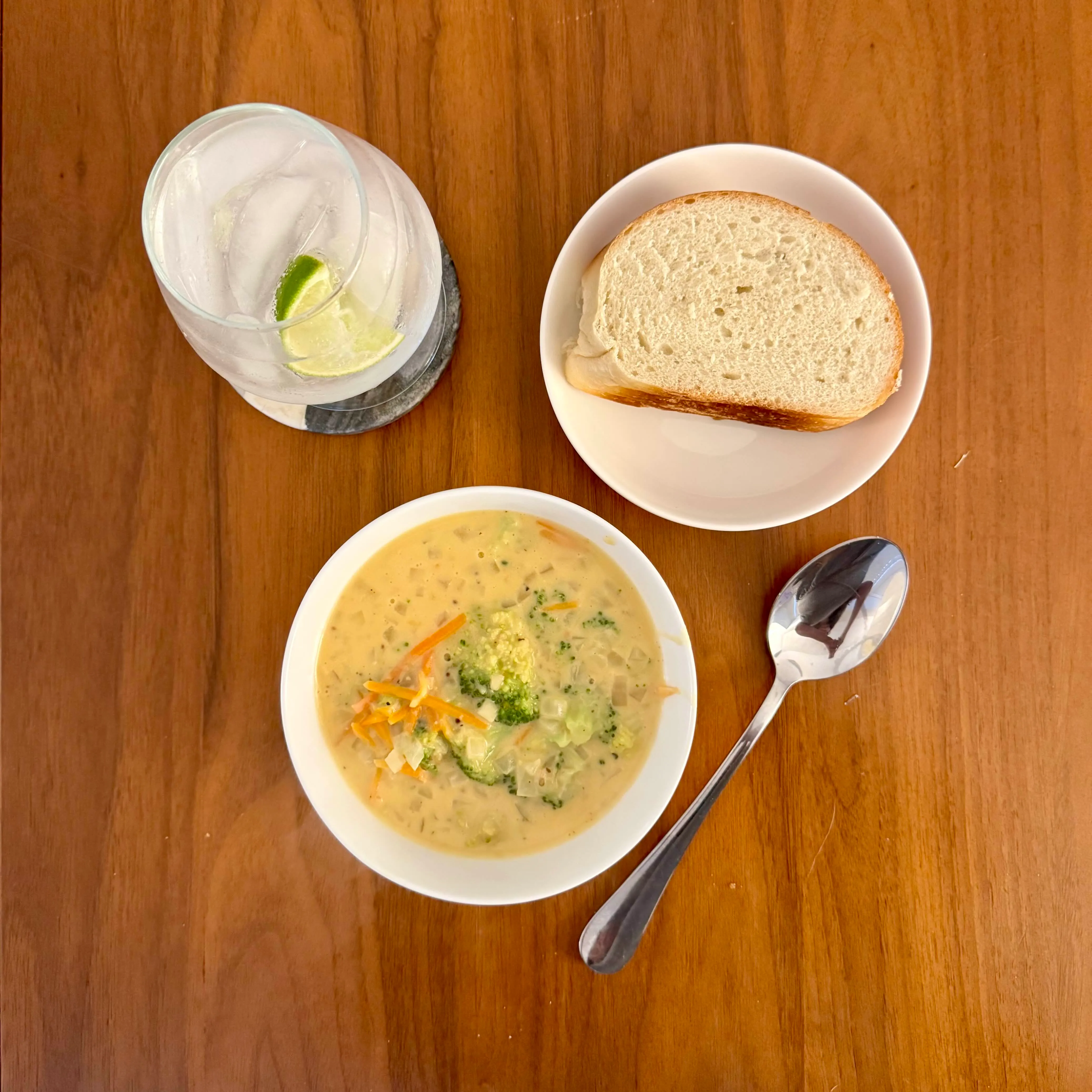 A bowl of broccoli-cheddar soup on a wood table, a glass of lime water and a thick slice of sourdough bread beside it, a long spoon resting on the right.