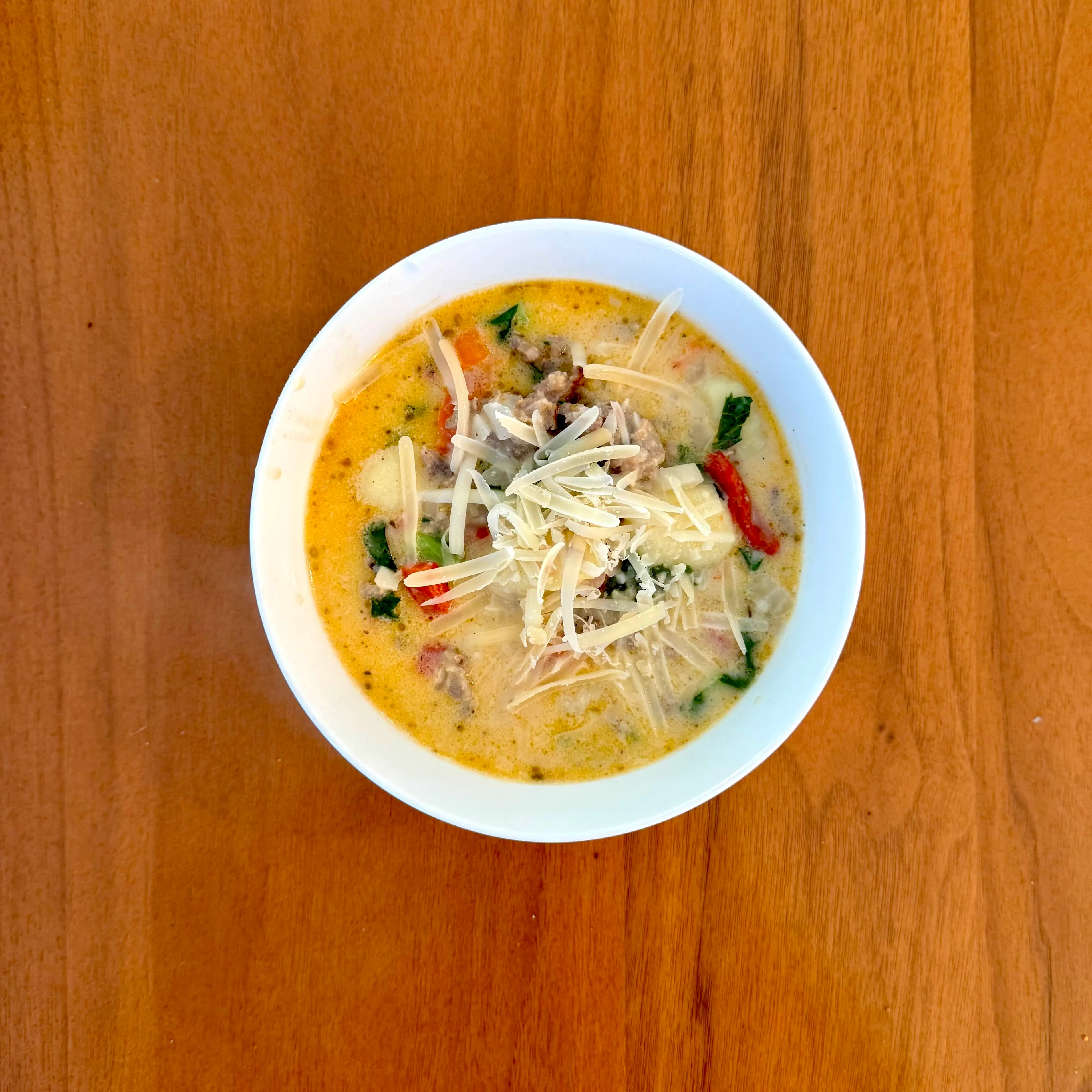 A white bowl of creamy sausage-and-greens soup with pasta and a scatter of shaved parmesan, flecked with bits of red pepper.