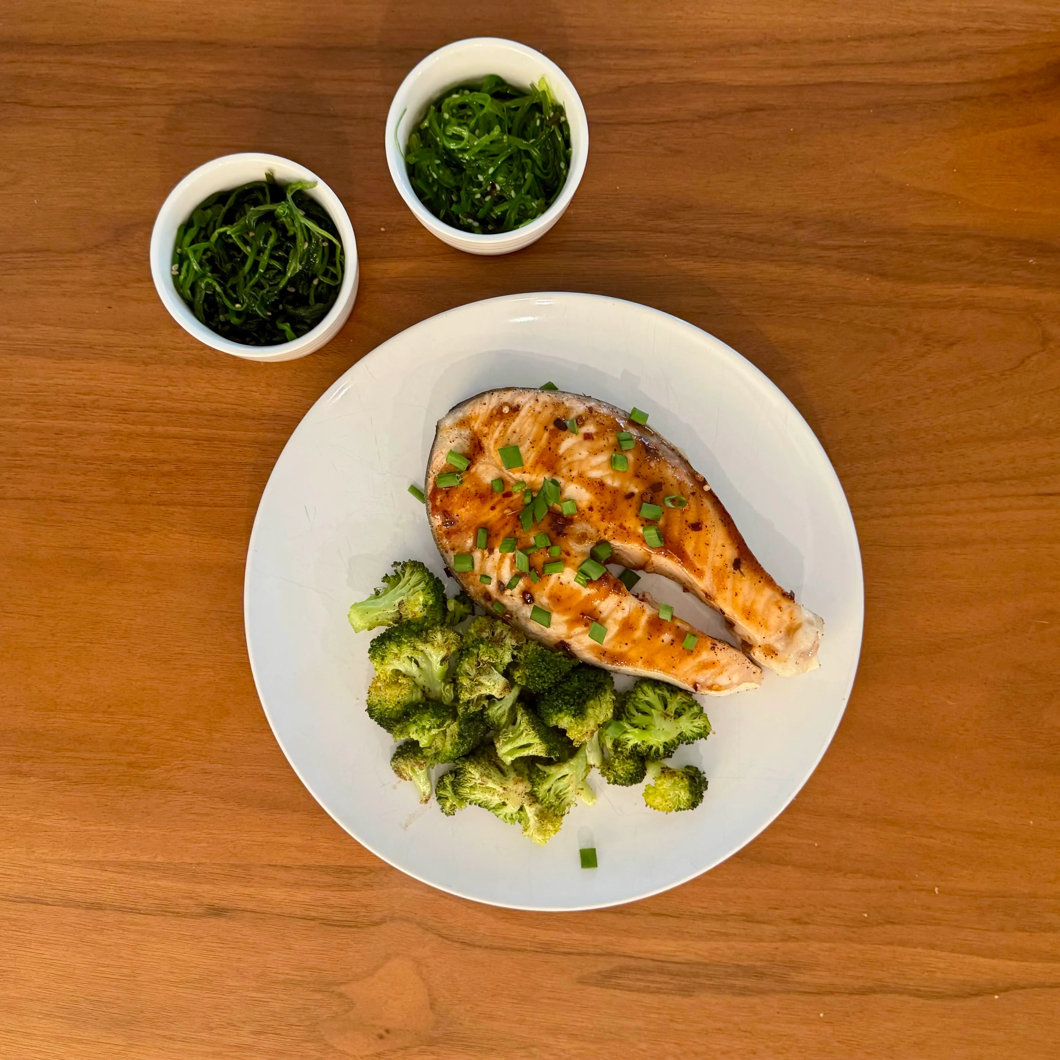 A salmon steak with a dark glaze and scallions, broccoli florets piled alongside, and two small ramekins of bright-green seaweed salad above the plate.