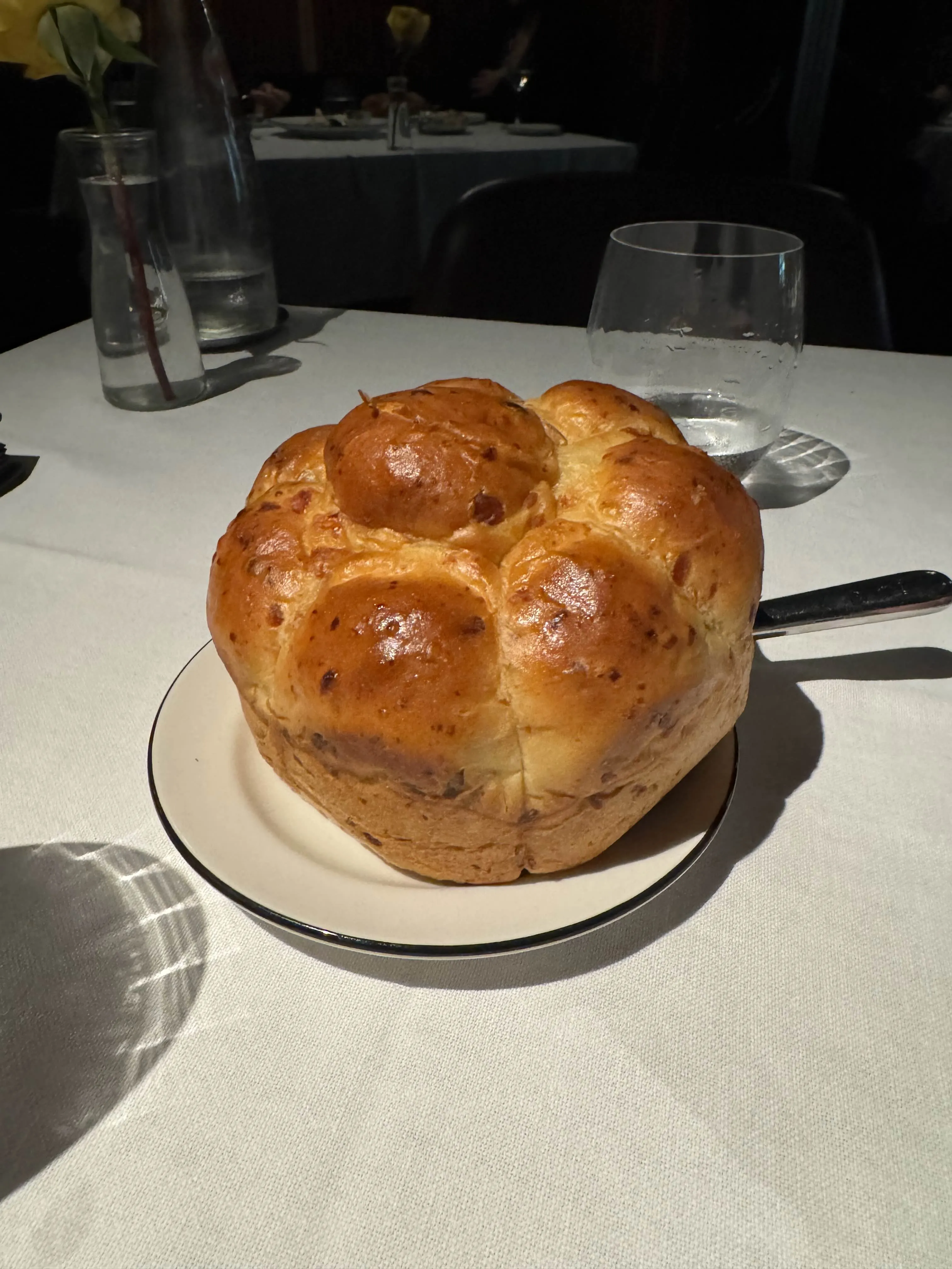 A round pull-apart bacon-and-cheese brioche loaf, golden-brown and glossy, served on a rimmed plate with a bread knife resting behind it on a white tablecloth.