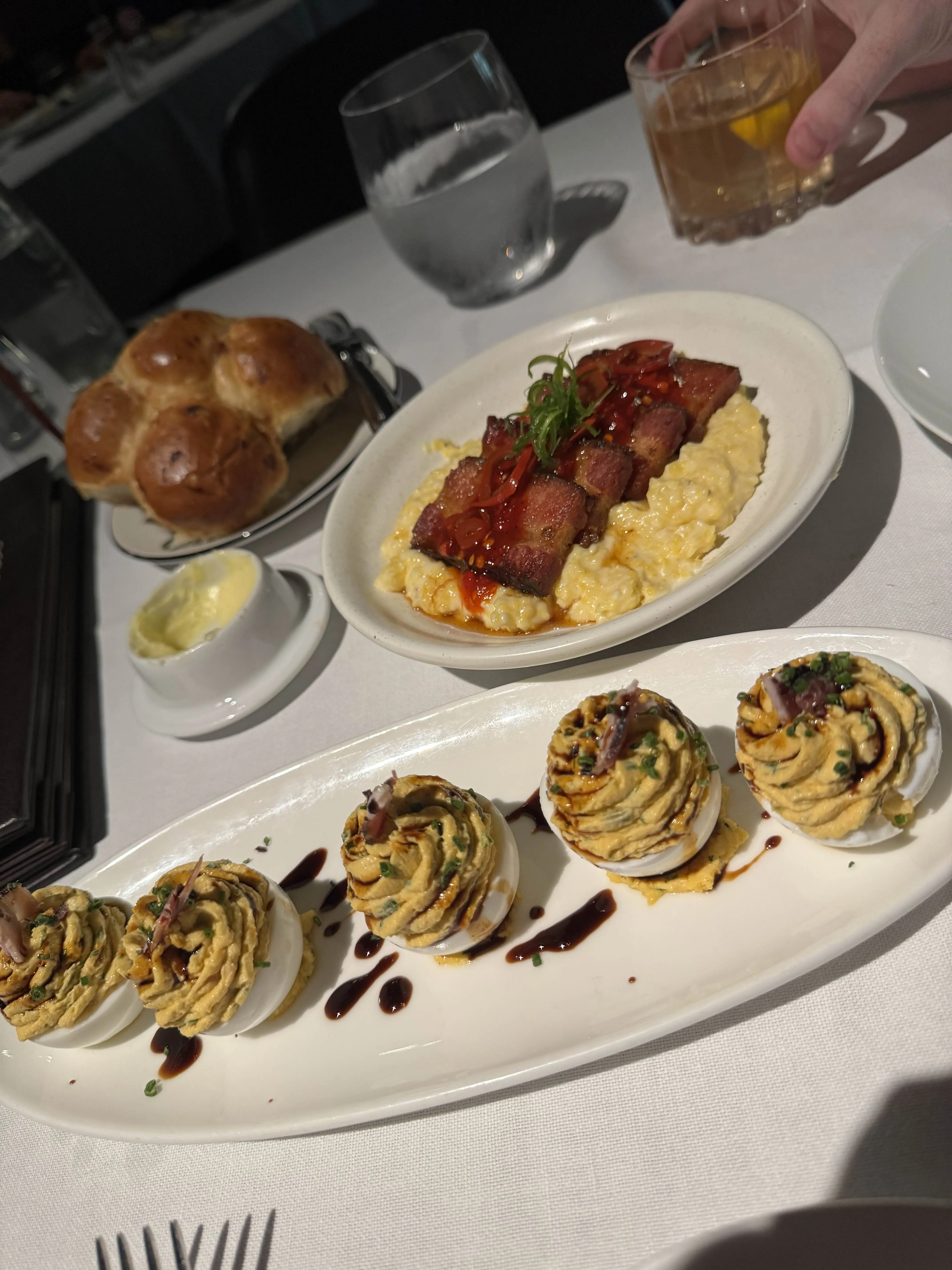 Deviled eggs with piped yolk and drizzled balsamic on a long white platter, a dish of lobster risotto with crisp bacon behind them, a whiskey cocktail in a rocks glass, and the brioche tucked to the side.