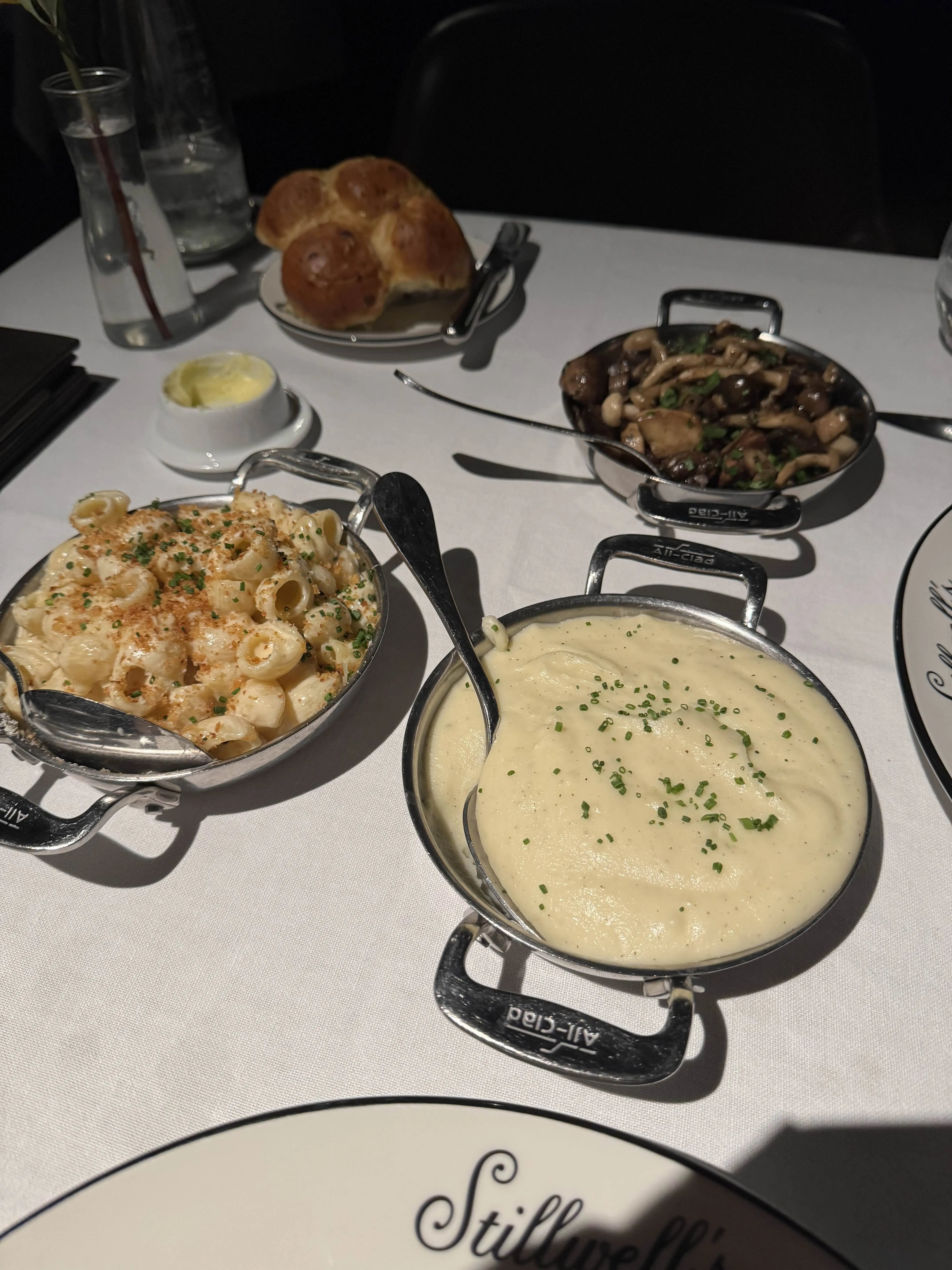 Three steakhouse sides in silver pans - pasta shells in bacon mac, creamy mashed with chives, and sautéed mushrooms - with a pat of butter and a small loaf of pull-apart bread behind.