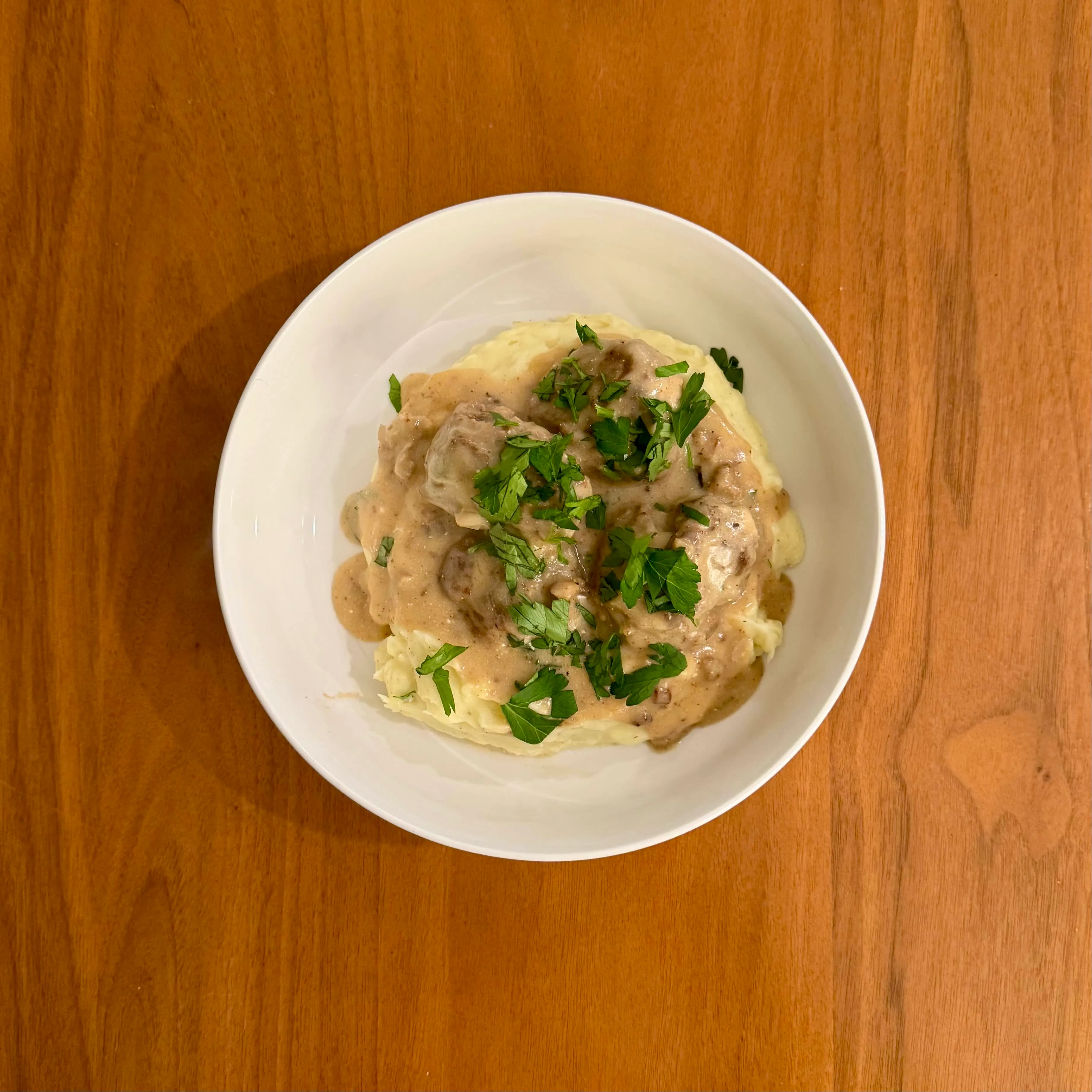 A mound of mashed potatoes covered in shredded chicken in a pale mushroom cream sauce and a generous scatter of fresh parsley.