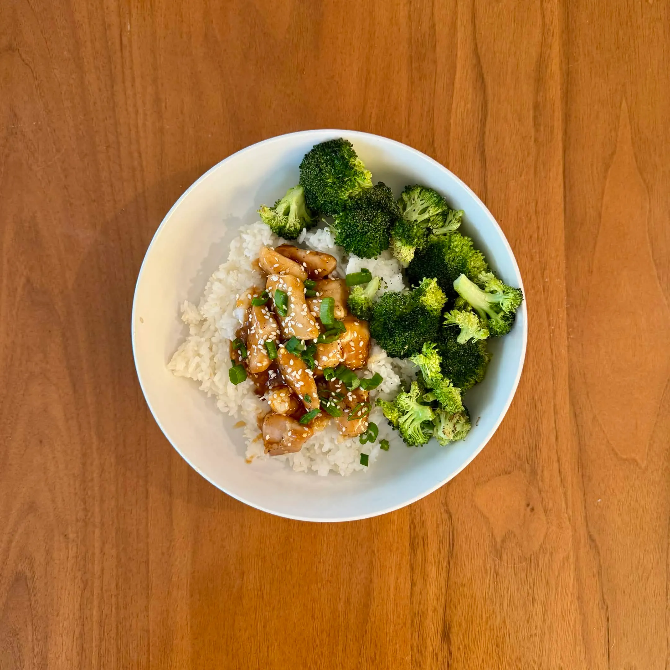 A bowl of white rice with teriyaki chicken, sesame seeds, chopped scallions, and a mound of roasted broccoli florets.