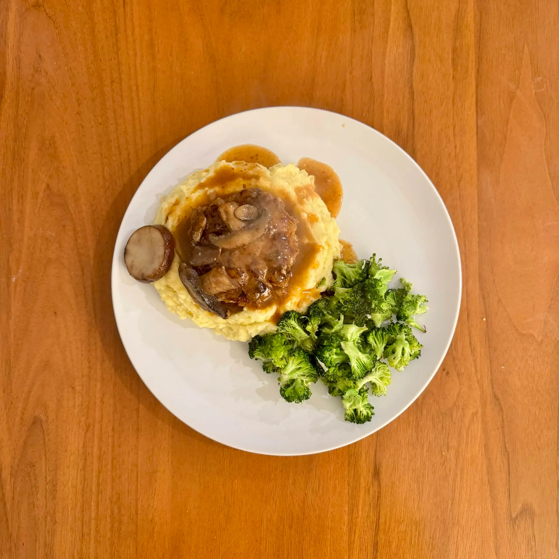 A plate with a salisbury steak and mushroom gravy over mashed potatoes, beside a pile of charred broccoli.
